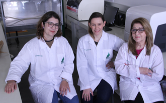 Foto no laboratório, com três mulheres sentadas em bancos, vestidas com jalecos