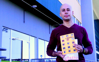 Foto do professor Igor em primeiro plano, mostrando dois exemplares da Revista Cidades, em frente ao bloco da Biblioteca.