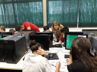 Em laboratório de informática, crianças estão em frente a computadores, e uma delas recebe a orientação de uma graduanda de Matemática