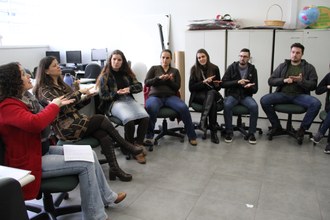 Foto em plano aberto mostra professoras e estudantes sentados em cadeiras, dispostos em formato circular, fazem sinais de Libras