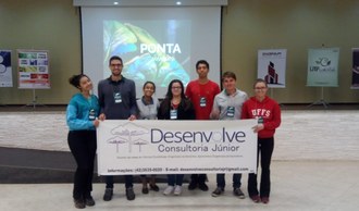 Na imagem cinco estudantes estão em pé segurando uma faixa que contém o logo da Desenvolve Consultoria Júnior e informações para contato