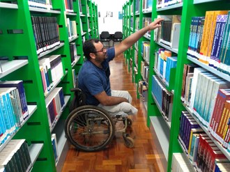 Na imagem um estudante cadeirante procura livros no acervo da biblioteca.