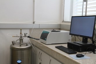 Na imagem observa-se o Calorímetro Diferencial de Varredura sobre uma bancada. Ao seu lado esquerdo um cilindro de gás acoplado ao equipamento e ao lado direito um computador, onde são apresentadas as análises realizadas pelo equipamento.