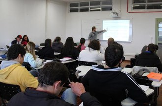 Na imagem um professor a frente da sala de aula aponta para conteúdo do quadro. Estudantes observam o docente.