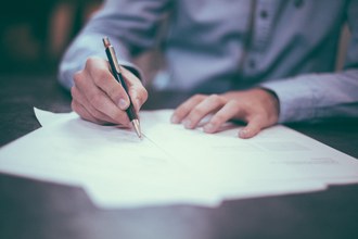 foto em que aparece as duas mãos de um homem sob um papel. Uma das mãos segura uma caneta apoiada no papel como se estivesse escrevendo.