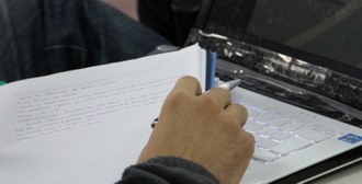 Foto, em plano fechado, de uma mão segurando um caderno e uma caneta