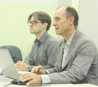 Dois homens, sentados lado a lado. Em primeiro plano, um homem segura um mouse de computador.