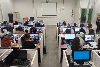 Foto de laboratório, com várias pessoas em baias, com fones de ouvido. Elas estão sentadas de costas, olhando para telas de computadores
