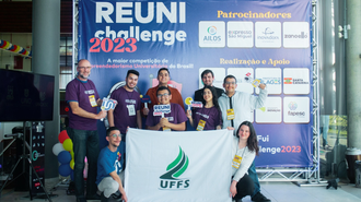 Imagem do grupo de estudantes e do professor. Parte da equipe está abaixada, segurando a bandeira da UFFS, e parte está em pé. Atrás deles, um painel do Reuni Challenge 2023
