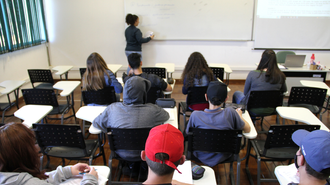 Foto de uma sala de aula. Os estudantes estão de costas, olhando para a professora e para o quadro, que estão, na foto, ao fundo.