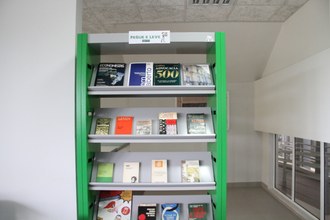 Foto em plano aberto da estante verde com materiais disponiveis para doação. No alto, uma plaquinha indicativa na qual está escrito "Pegue e Leve", com uma imagem de uma pessoa com livros na mão