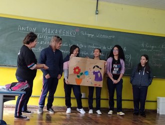 Fotografia mostra seis estudantes em pé dentro de uma sala de aula. Duas estudantes estão no centro da foto segurando um cartaz. Atrás dos estudantes uma lousa com várias anotações.