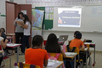 Crianças estudando em uma sala de aula. Ao fundo, professora auxilia aluna com atividade.
