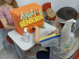 Criança com olhos vendados, colocando a mão em uma caixa. Na caixa está escrito análise sensorial.